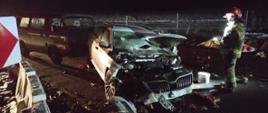 Na zdjęciu wrak rozbitego samochodu osobowego za nim samochód osobowy typu bus który się z nim zderzył. Po prawej strażak PSP w ubraniu specjalnym i hełmie z włączoną latarką w ręku. Na jezdni porozrzucane części samochodu. W tle zaśnieżone pole.