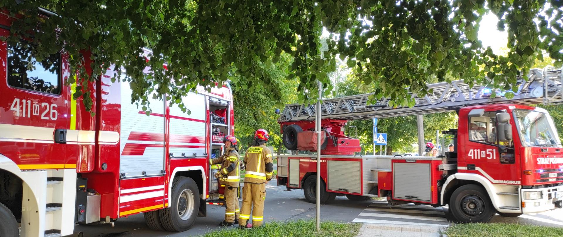 Z lewej strony duży czerwony samochód strażacki i przy nim dwóch strażaków. Z prawej strażacka drabina na samochodzie.