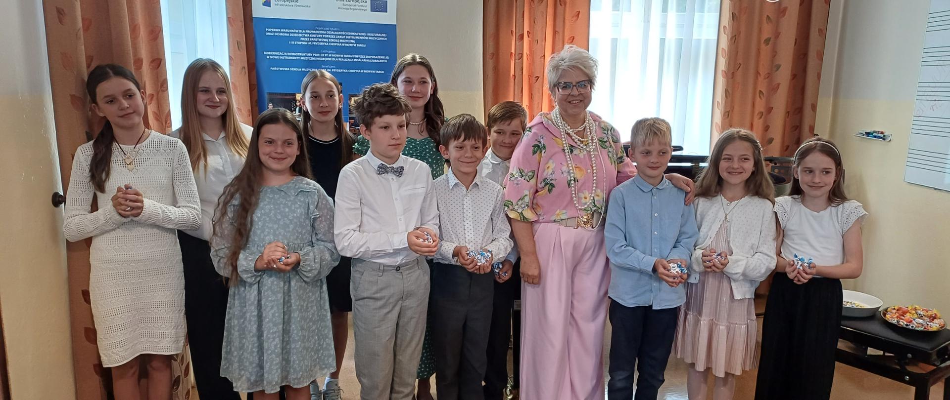 Zdjęcie przedstawia grupę około dwunastu osób, w większości dzieci i młodzieży, stojących obok starszej kobiety w pomieszczeniu przypominającym salę muzyczną lub lekcyjną. Osoby te stoją w półkolu lub luźnej linii, uśmiechając się do aparatu.
Kobieta w centrum:
Starsza kobieta w jasnych, siwych włosach, krótkich i puszystych. Ubrana jest w luźną, kwiecistą bluzkę w odcieniach różu, zieleni i bieli, oraz jasne, różowe spodnie. Na szyi ma naszyjnik z dużych, okrągłych pereł. Stoi uśmiechnięta, z rękami ułożonymi z przodu. Wydaje się być nauczycielką lub opiekunką grupy.
Dzieci i młodzież (od lewej do prawej):
Pierwsza osoba z lewej: Młoda dziewczyna w jasnej, prawdopodobnie kremowej lub białej sukience z długimi rękawami. Ma długie, ciemne włosy spięte w kitkę. Uśmiecha się.
Druga osoba z lewej: Dziewczyna w ciemnej spódnicy i jasnej bluzce z długim rękawem. Ma długie, proste, jasne włosy.
Trzecia osoba z lewej (z przodu): Dziewczynka w jasnej, niebieskawej sukience z krótkim rękawem, ozdobionej delikatnym wzorem. Ma ciemne włosy, rozpuszczone. Trzyma w rękach mały, jasnoniebieski przedmiot.
Czwarta osoba z lewej (za trzecią): Dziewczyna w ciemnej sukience, częściowo zasłonięta.
Piąta osoba z lewej (chłopiec): Chłopiec w białej koszuli, szarych spodniach i muszce. Ma jasne włosy. Trzyma w rękach mały, biały przedmiot.
Szósta osoba z lewej (chłopiec): Chłopiec w białej koszuli w ciemne kropki i ciemnych spodniach. Ma krótkie, jasne włosy. Trzyma w rękach mały, jasnoniebieski przedmiot.
Siódma osoba z lewej (chłopiec, za szóstym): Chłopiec w jasnej koszuli, stojący częściowo za inną osobą.
Ósma osoba (chłopiec, obok starszej kobiety): Chłopiec w jasnoniebieskiej koszuli i ciemnych spodniach. Ma jasne włosy. Trzyma w rękach mały, jasnoniebieski przedmiot.
Dziewiąta osoba (dziewczynka): Dziewczynka w jasnej, różowej, plisowanej spódnicy i jasnym sweterku. Ma długie, jasne włosy. Trzyma w rękach mały, jasnoniebieski przedmiot.
Dziesiąta osoba (dziewczynka): Dziewczynka w ciemnej spódnicy i jasnej bluzce z falbankami. Ma długie, jasne włosy spięte w kitkę.
Tło:
Po lewej stronie tła widoczna jest duża, pionowa plansza informacyjna w odcieniach niebieskiego i zielonego z logotypami Funduszy Europejskich i Unii Europejskiej. Na planszy znajdują się zdjęcia fortepianów i innych instrumentów muzycznych. W centralnej i prawej części tła widoczna jest ściana w jasnym, beżowym kolorze oraz ciemne, brązowe zasłony z delikatnym wzorem. Po lewej stronie, na dole, widoczny jest kaloryfer. Po prawej stronie, na dole, znajduje się fragment mebla, prawdopodobnie taboretu fortepianowego, z tacą z drobnymi słodyczami lub upominkami. Całość sugeruje atmosferę uroczystości lub podsumowania osiągnięć, prawdopodobnie po koncercie lub konkursie muzycznym.