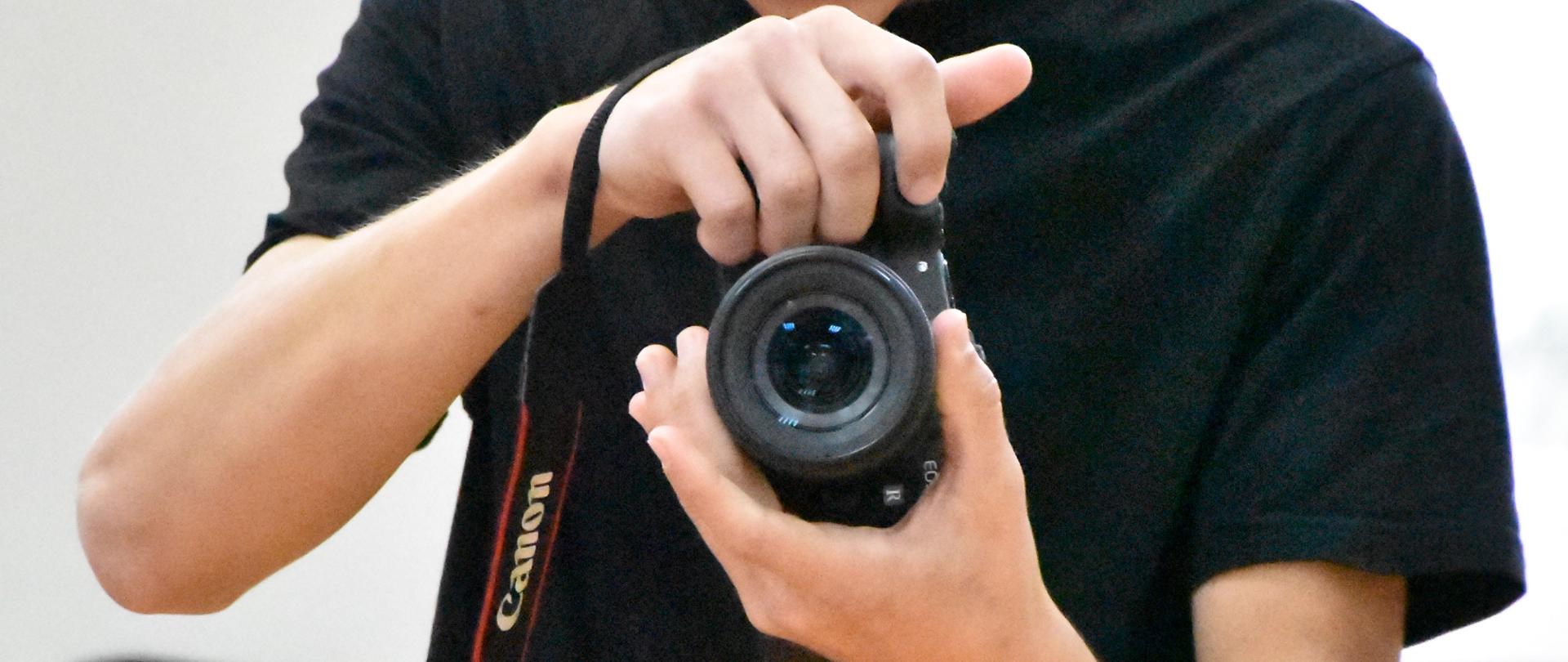 Młody mężczyzna trzyma w dłoniach aparat fotograficzny marki Canon i uważnie przegląda wykonane zdjęcia na ekranie podglądu. Wygląda na osobę dokumentującą wydarzenie lub pracującą jako fotograf