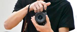 Młody mężczyzna trzyma w dłoniach aparat fotograficzny marki Canon i uważnie przegląda wykonane zdjęcia na ekranie podglądu. Wygląda na osobę dokumentującą wydarzenie lub pracującą jako fotograf.