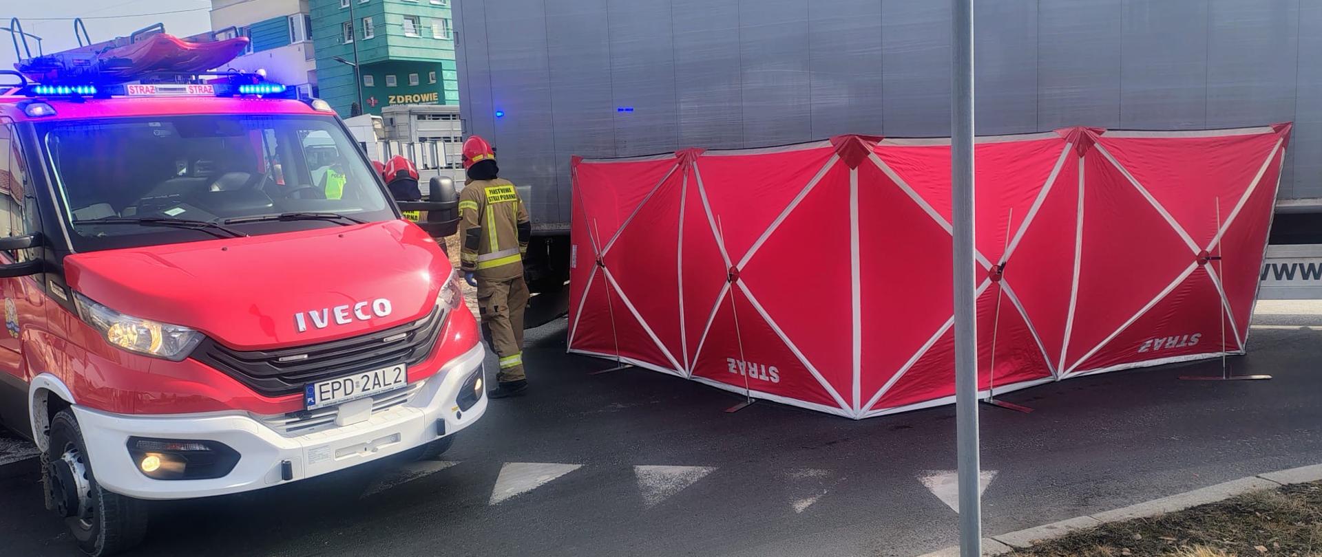 Czerwony parawan rozłożony przy samochodzie ciążowym obok stoją strażacy i czerwony samochód strażacki 