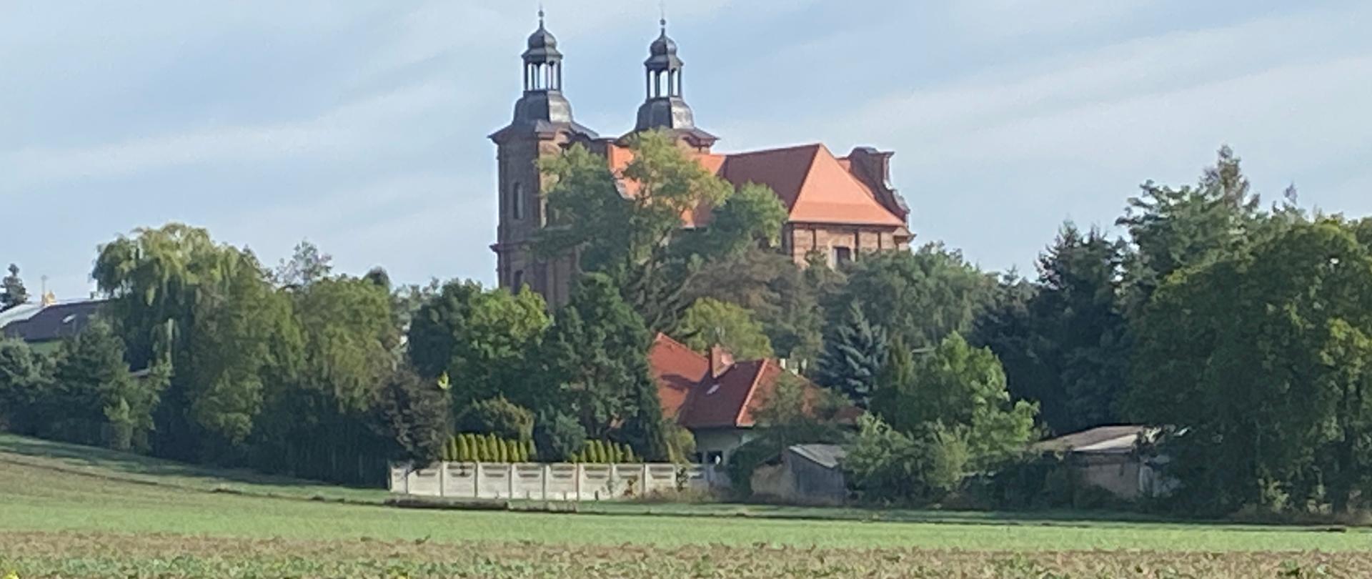 Zdjęcie przedstawiające miejscowość Lądek. W środkowej części nad horyzontem znajduje się kościół parafii p.w. Św. Mikołaja Biskupa w Lądku, poniżej pola i kwiaty.