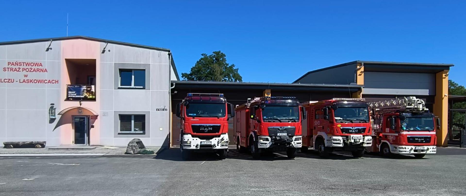 Zdjęcie przedstawia budynek Jednostki Ratowniczo-Gaśniczej Państwowej Straży Pożarnej w Jelczu-Laskowicach. Przed bramami garażowymi, na asfaltowym placu manewrowym, stoją zaparkowane cztery czerwone pojazdy pożarnicze.
Od lewej strony widoczne są: trzy samochody ratowniczo-gaśnicze (marki MAN) oraz samochód specjalny z drabiną mechaniczną lub podnośnikiem hydraulicznym (również marki MAN). Po lewej stronie kadru znajduje się dwukondygnacyjna, jasnoszara część biurowo-socjalna budynku z czerwonym napisem „PAŃSTWOWA STRAŻ POŻARNA W JELCZU-LASKOWICACH”. Zdjęcie zostało wykonane przy bezchmurnym, niebieskim niebie.