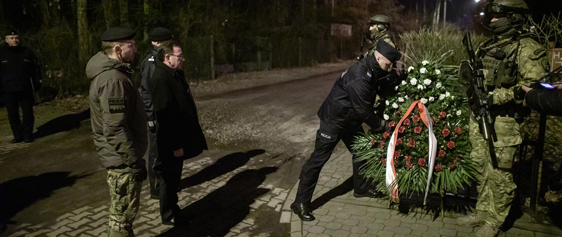 Na zdjęciu widać Mariusza Kamińskiego ministra SWiA wraz z gen. insp. Jarosławem Szymczykiem komendantem głównym Policji oraz mł. insp. Dariuszem Ziębą dowódcą Centralnego Pododdziału Kontrterrorystycznego Policji BOA stojących przed pomnikiem upamiętniającym poległych policjantów. Policjant im towarzyszący kładzie wieniec. Widać również dwóch policjantów Centralnego Pododdziału Kontrterrorystycznego Policji BOA stojących na posterunku przy pomniku.