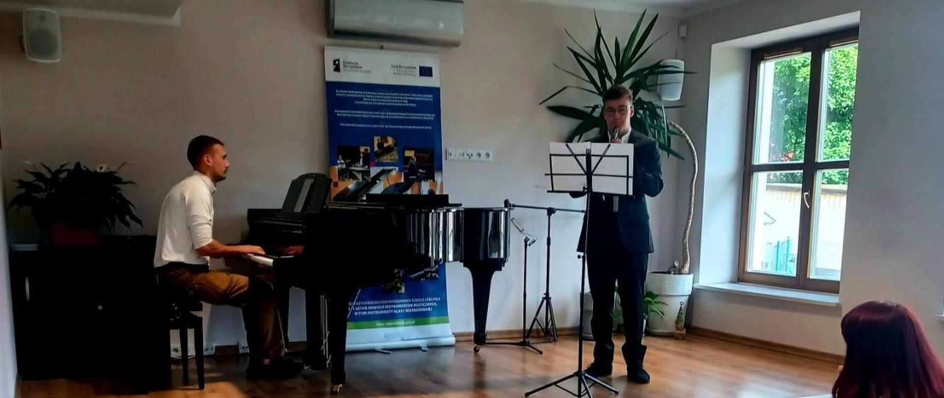 Sala kameralna, na scenie od prawej strony stoi pulpit, kwiatki, chłopiec, który gra na oboju. Następnie na czarnym fortepianie gra pan akompaniator. Za fortepianem baner reklamowy zakupionych instrumentów z funduszu Unii Europejskiej. Pod sceną widownia siedzi na krzesłach.