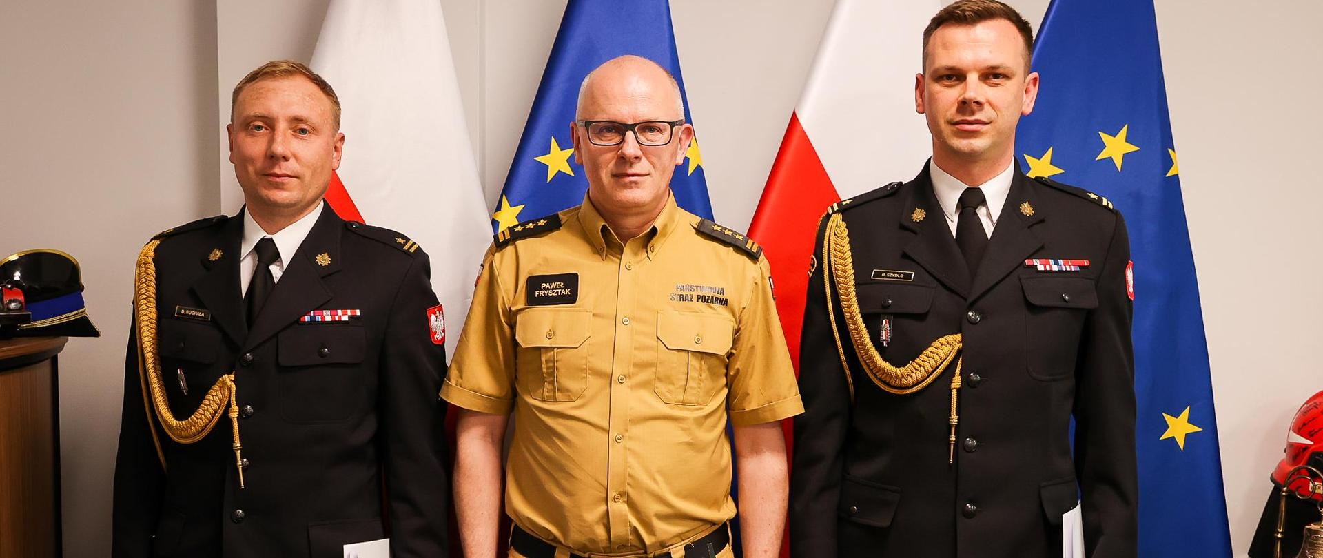 Biro komendanta głównego PSP. Na tle unijnych i biało-czerwonych flag – trzy osoby – strażacy w umundurowaniu wyjściowym i służbowym. Od lewej mł.bryg. Dariusz Ruchała, st.bryg. Paweł Frysztak, mł.bryg. Bogusław Szydło. 