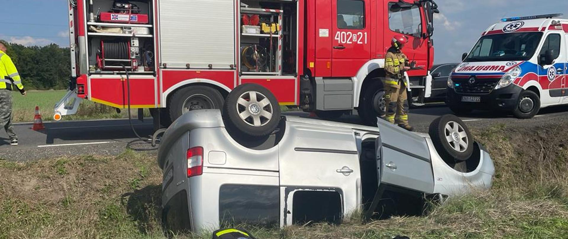 Wypadek samochodu osobowego na DW 385 w pobliżu miejscowości Grodków - zdjęcie przedstawia samochód osobowy leżący na dachu w przydrożnym rowie, w tle ustawiony jest pojazd pożarniczy oraz karetka pogotowia