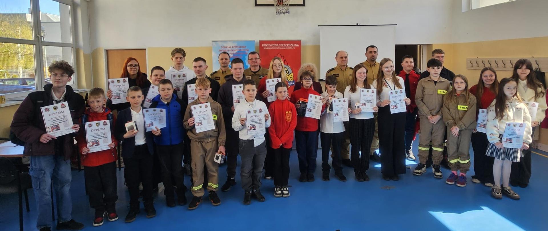 Na zdjęciu widać grupę dzieci i dorosłych ustawionych w rzędzie w sali gimnastycznej, pod koszem do koszykówki. Wiele osób trzyma w rękach dyplomy, a za nimi stoją osoby w mundurach. Z lewej strony znajdują się duże przeszklenia okienne, z prawej strony drzwi i wieszaki na ścianie. Za osobami pozującymi do zdjęcia znajdują się dwa kolorowe banery.