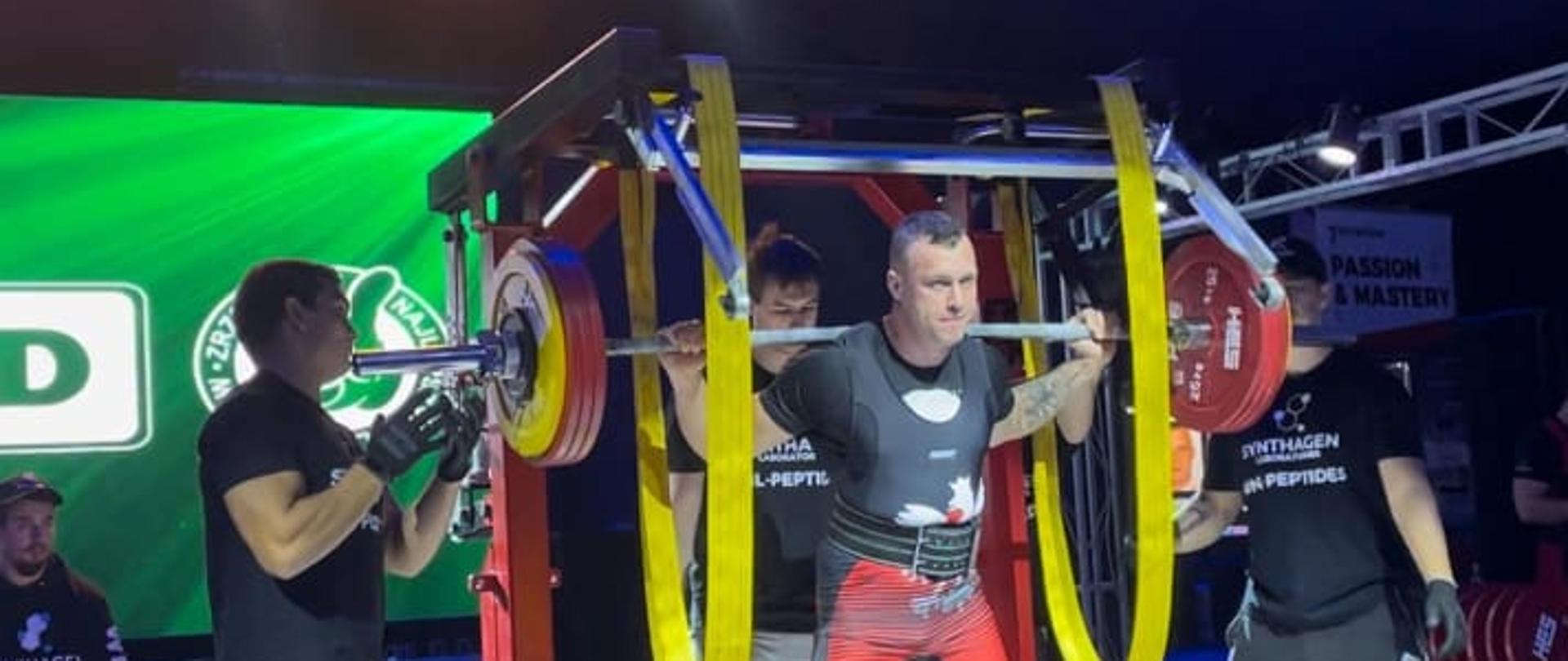 Mężczyzna stoi na podwyższeniu na plecach dźwiga sztangę kulturystyczną.