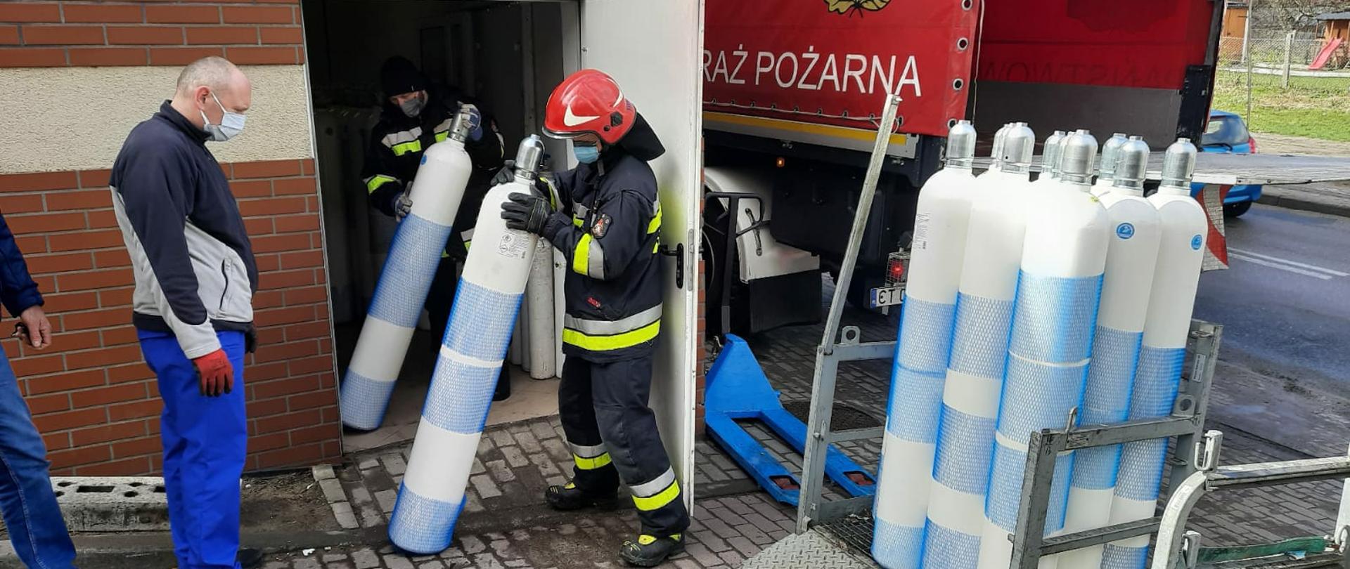 Na zdjęciu widać dwóch strażaków trzymających butle tlenowe, stojącego obok nich mężczyznę, nogę jeszcze jednej osoby oraz kilka butli tlenowych w skrzyni transportowej. Tłem zdjęcia jest budynek oraz fragment samochodu cięzarowego z napisem straż pożarna.
