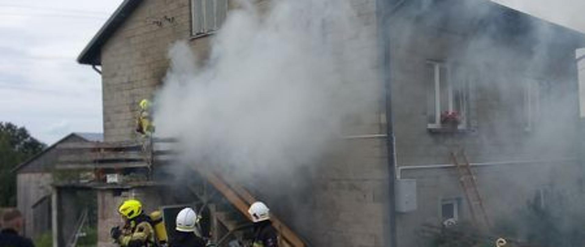 Strażacy w trakcie działań w Kapałowie, trzech przed budynkiem, jeden widoczny na schodach zewnętrznych wchodzi w zadymienie do budynku, z drzwi wydobywa się dużo białego dymu