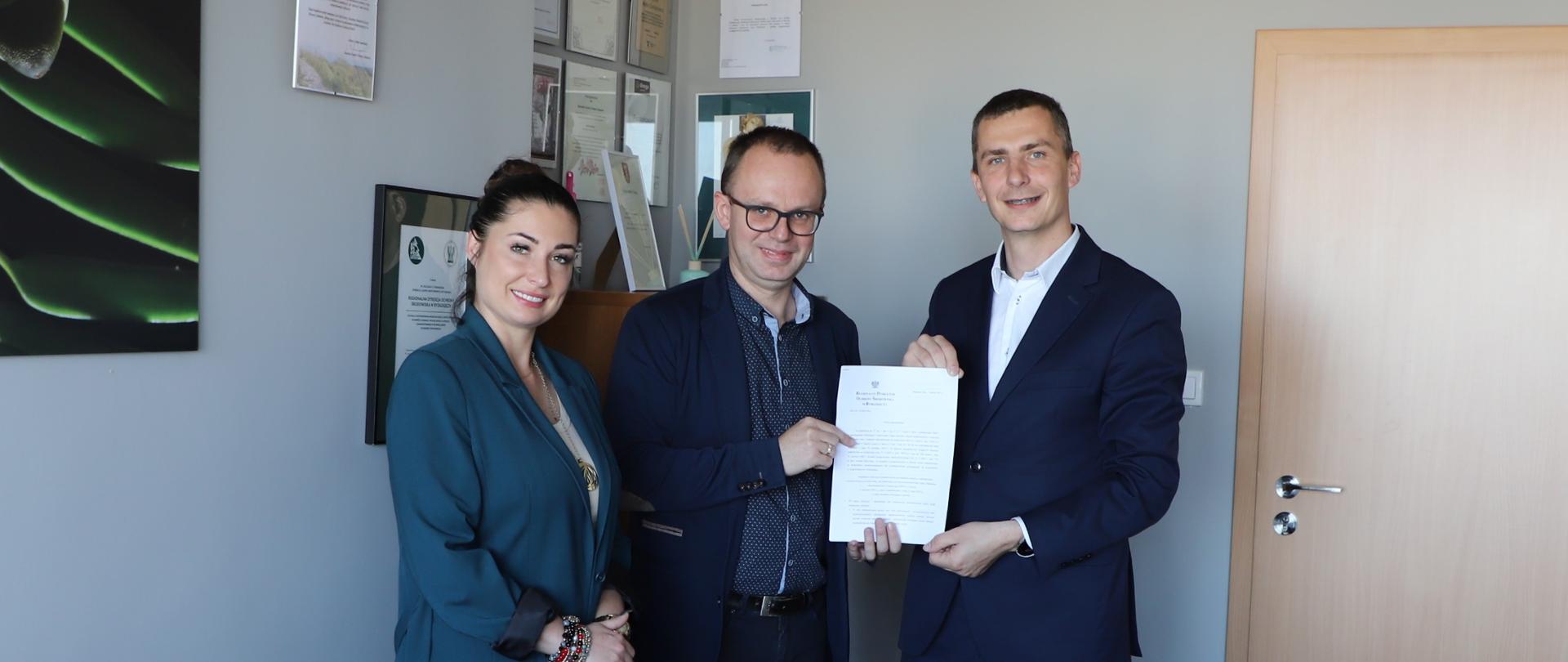 Pan Szymon Kosmalski, Regionalny Dyrektor Ochrony Środowiska w Bydgoszczy w środku, wręcza dokument Panu Krzysztofowi Kukuckiemu, Zastępcy Prezydenta Włocławka po prawej. Po lewej stronie stoi Pani Magdalena Nowak, Dyrektor Biura Poselskiego Minister Anny Gembickiej.