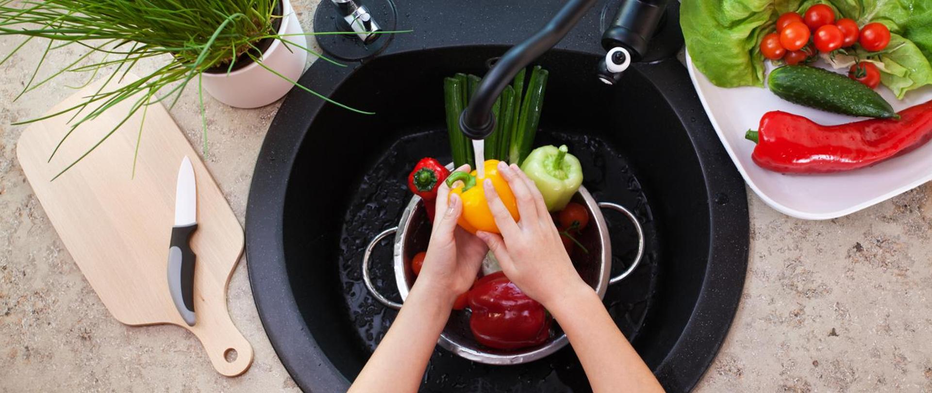 Na zdjęciu widoczne dłonie myjące owoce i warzywa pod bieżącą wodą w zlewie kuchennym.