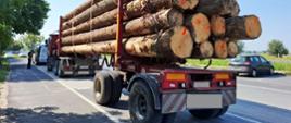 Ciężarówka z przyczepą transportującą długie kłody drewna ustawione warstwowo, zaparkowana przy drodze, z widocznymi oznaczeniami ostrzegawczymi.