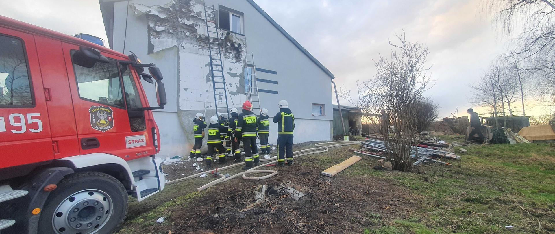 Zdjęcie wykonane w godzinach popołudniowych. Na zdjęciu widoczni strażacy , którzy ustawiają drabiny przystawne o budynek jednorodzinny . W tle widoczny samochód pożarniczy.