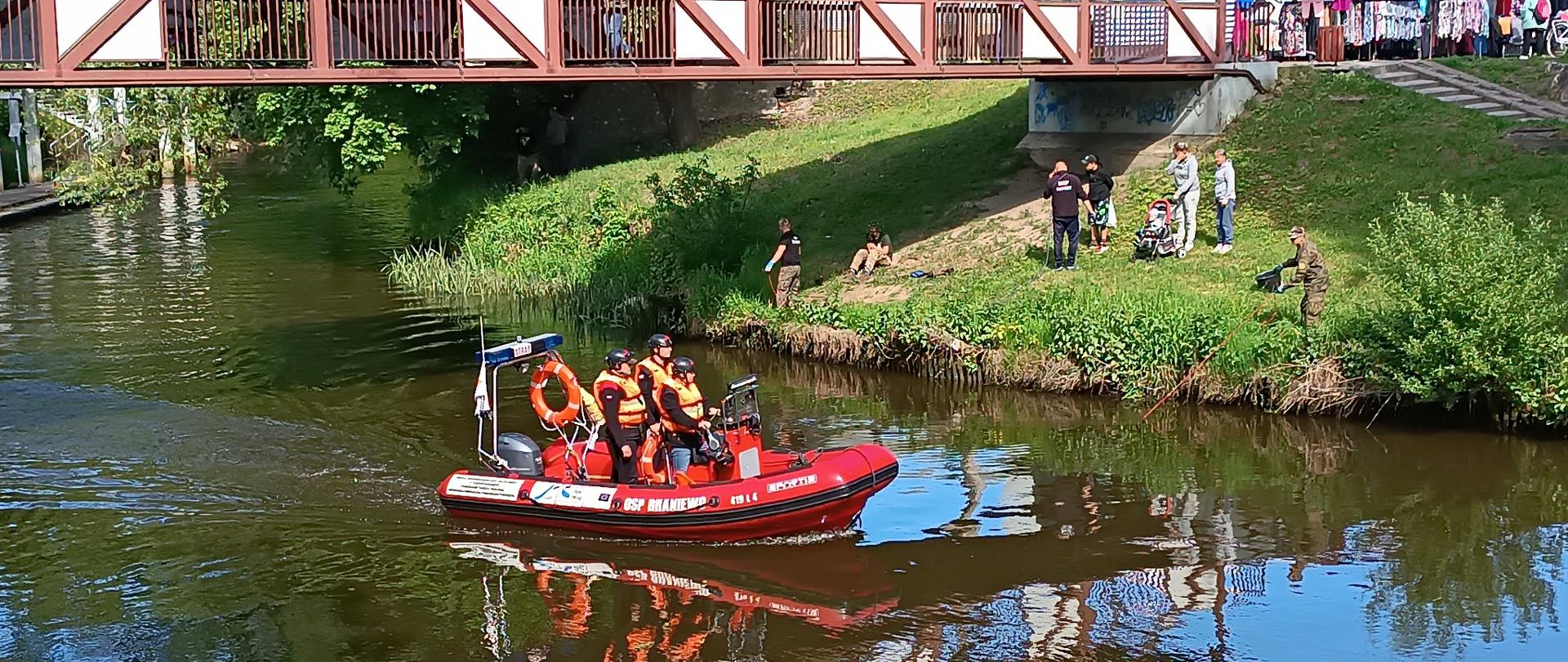 Czerwona lódź motorowa na wodzie, płynie wzdłuż rzeki nad mostem. W łodzi trzech strażaków.