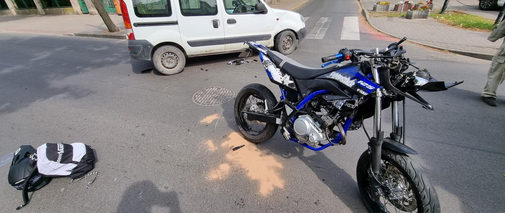 Na pierwszym planie motocykl (czarno-niebieski) i samochód osobowy koloru białego po zderzeniu na skrzyżowaniu 2 ulic. Po lewej stronie leży plecak i kask motocyklisty