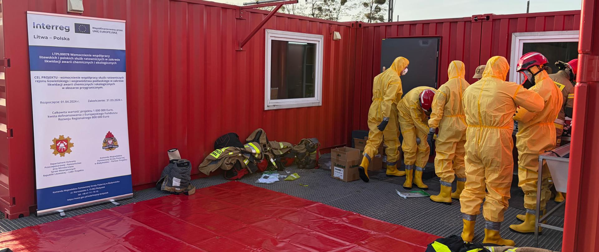Projekt LTPL00078 "Wzmocnienie współpracy litewskich i polskich służb ratowniczych w zakresie likwidacji awarii chemicznych i ekologicznych" - strażacy w trakcie szkolenia z zakresu dekontaminacji i ratownictwa chemiczno-ekologicznego, po lewej stronie baner projektowy. Na dole rozłożona czerwona mata. 