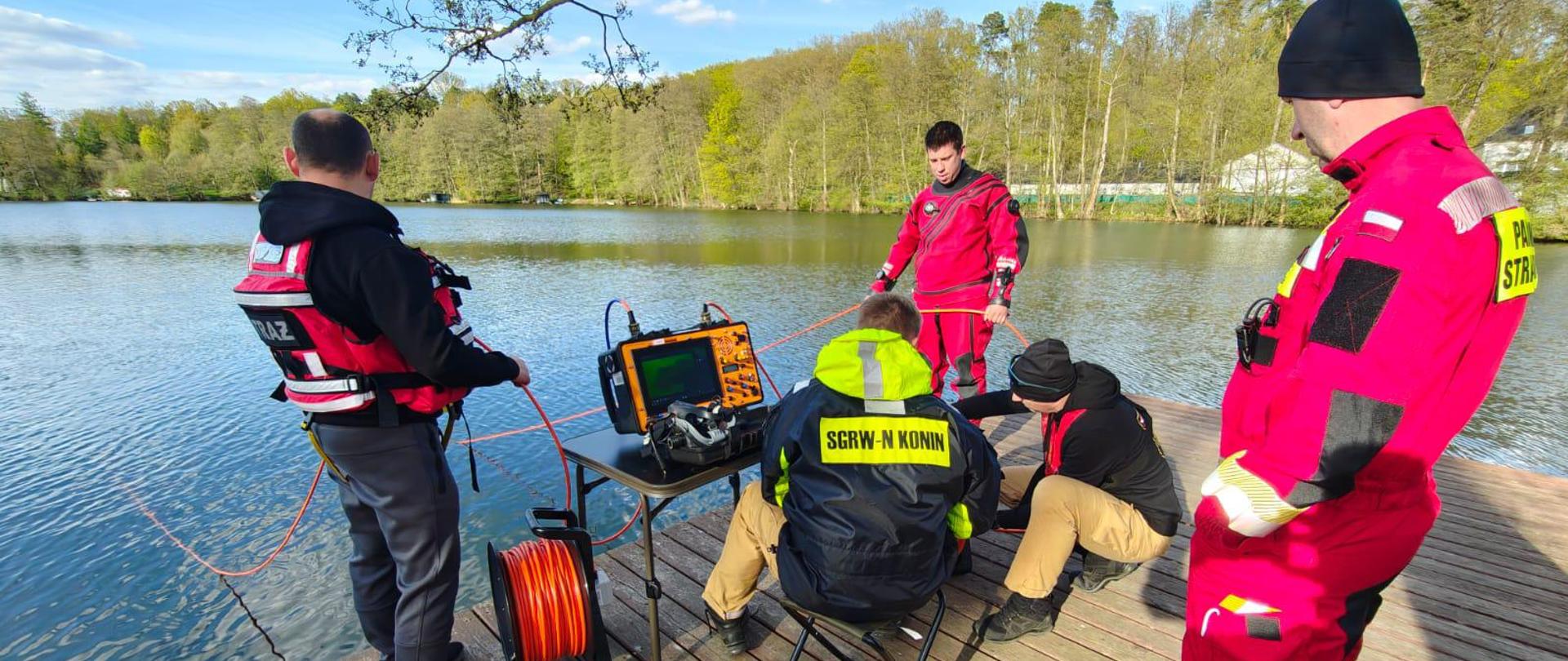 Strażacy nurkowie przygotowują się do zejścia pod wodę, stojąc na pomoście przy jeziorze.