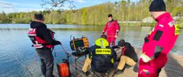 Strażacy nurkowie przygotowują się do zejścia pod wodę, stojąc na pomoście przy jeziorze.