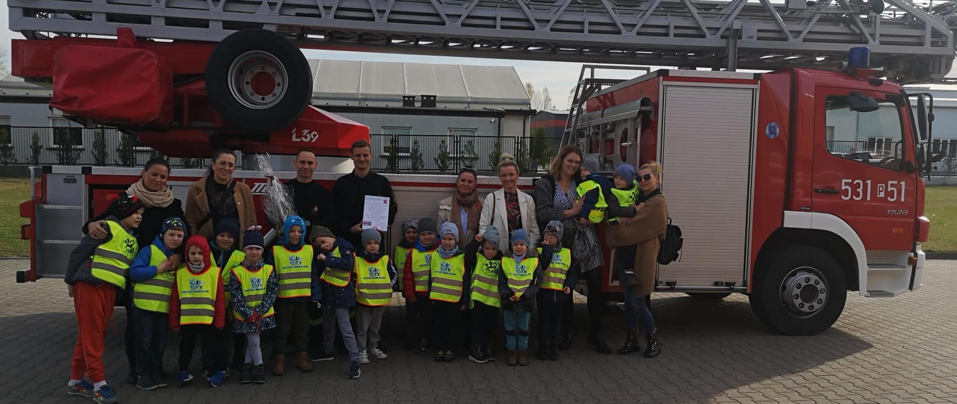 Przedszkolaki z opiekunami oraz dwóch strażaków stojących na tle samochodu-drabiny pożarniczej.