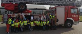 Przedszkolaki z opiekunami oraz dwóch strażaków stojących na tle samochodu-drabiny pożarniczej.
