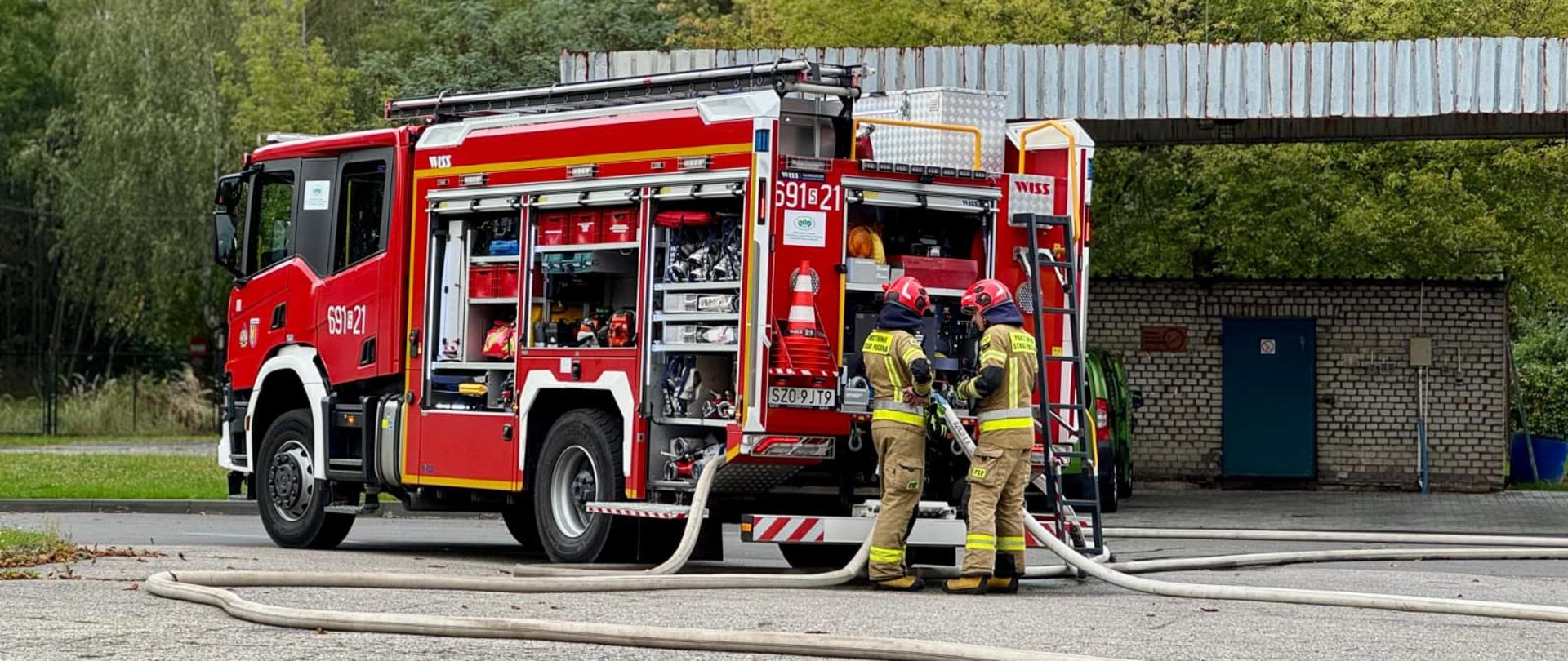 wóz strażacki z otwartymi bocznymi schowkami, w których znajdują się różne narzędzia i sprzęt gaśniczy. Dwóch strażaków w pełnym umundurowaniu stoi z tyłu pojazdu, trzymając wąż strażacki podłączony do auta. Wąż rozciąga się po asfalcie. W tle widoczne są drzewa, mały ceglany budynek z niebieskimi drzwiami oraz metalowa wiata.