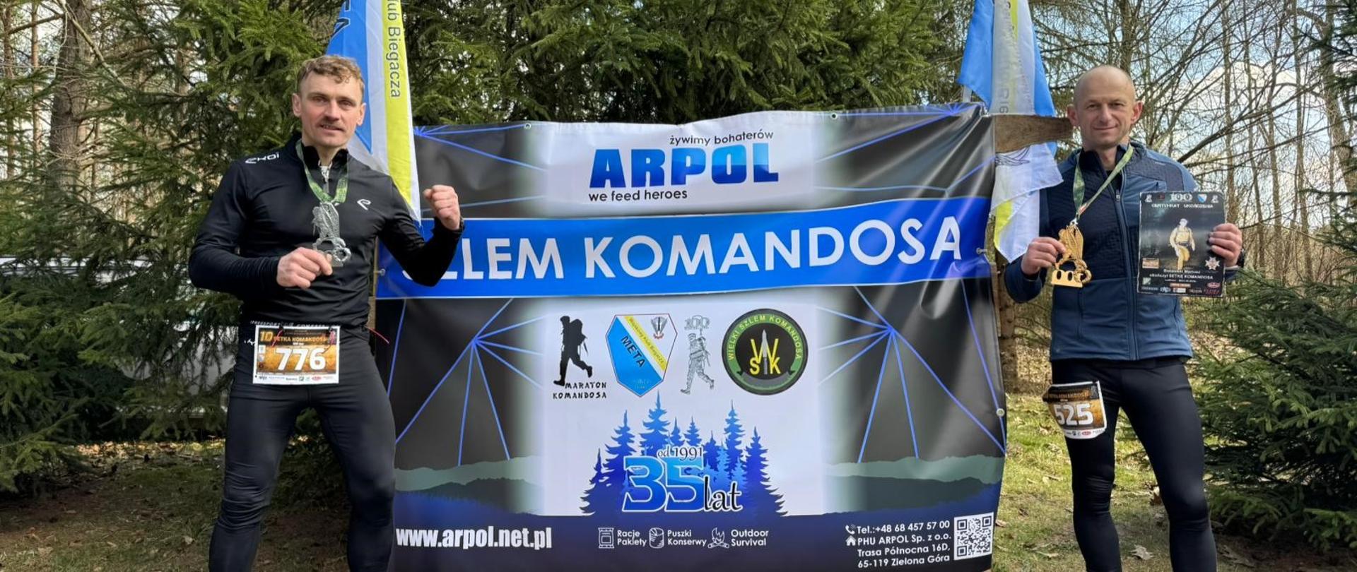 Dwóch mężczyzn w strojach sportowych pozuje do zdjęcia z dyplomem i medalem na tle biało-niebieskiej ścianki z napisem „SZLEM KOMANDOSA”.