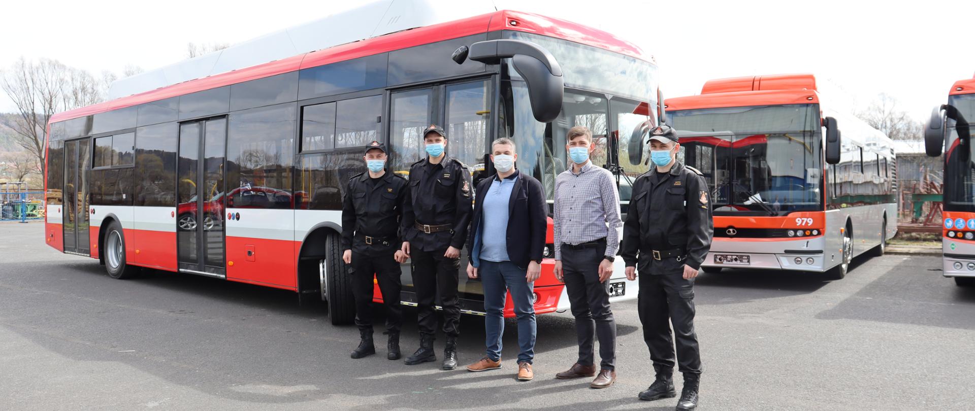 Zdjęcie przedstawia 5 osób - 3 funkcjonariuszy PSP oraz 2 pracowników firmy Autosan. Osoby stoją przed pojazdem - autobusem koloru czerwono - biało - czarnego. W tle zdjecia widać inne autobusy.