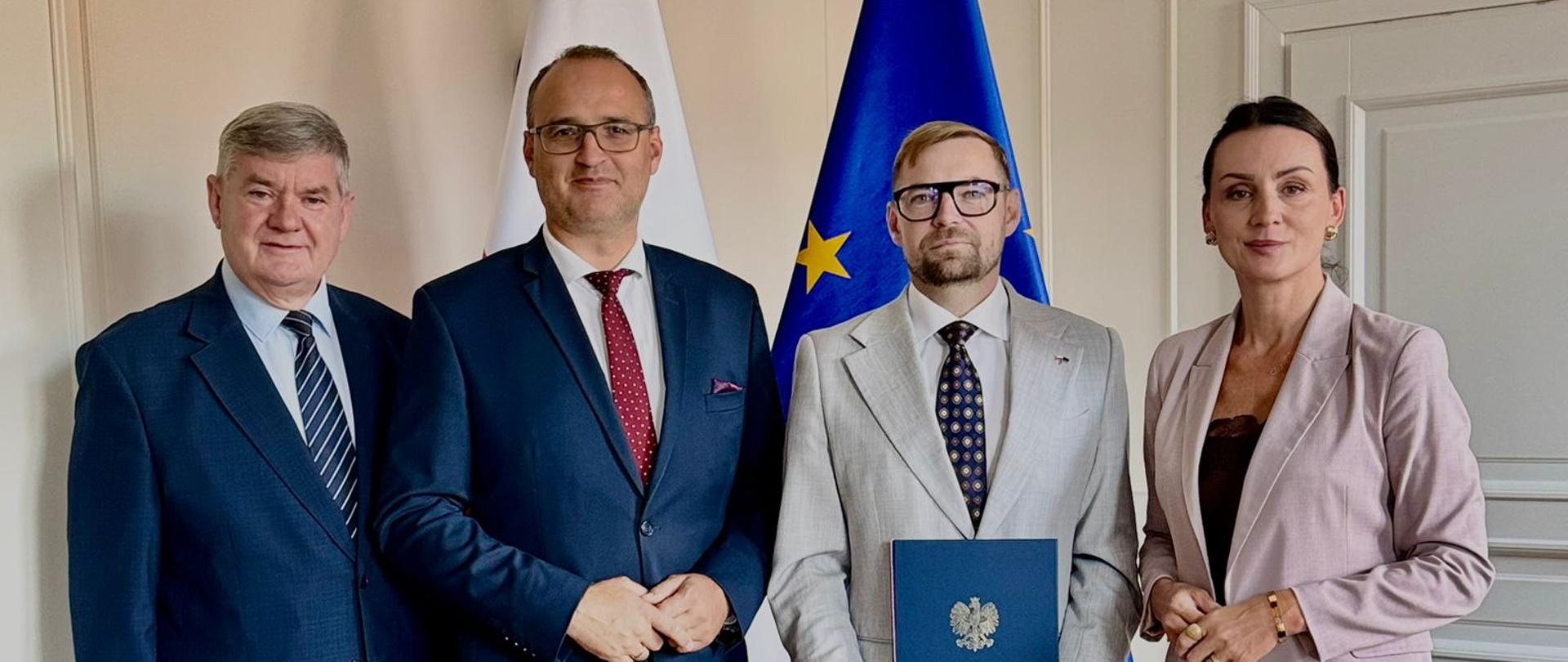 Cztery osoby stoją obok siebie w jasnym pomieszczeniu, na tle flag Polski i Unii Europejskiej. Od lewej: mężczyzna w ciemnym garniturze i niebieskim krawacie, drugi mężczyzna w ciemnym garniturze i bordowym krawacie w kropki, trzeci mężczyzna w jasnym, popielatym garniturze, okularach i wzorzystym krawacie, trzymający w dłoniach niebieską teczkę z godłem Polski, oraz stojąca po prawej stronie kobieta w jasnoróżowym garniturze i ciemnej bluzce. Wszyscy patrzą w stronę aparatu.