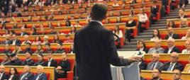 mężczyzna w ciemnym garniturze, wicewojewoda, stoi na mównicy tyłem w tle widać uczestników wydarzenia na widowni