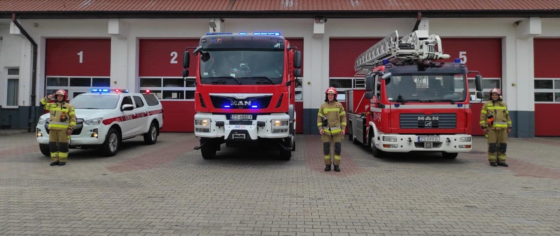 Przed strażnicą stoją trzy samochody oraz trzech strażaków oddających hołd powstańcom warszawskim.