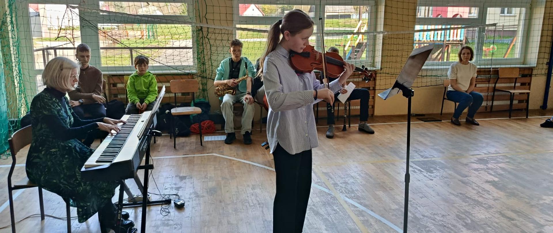 Młoda dziewczyna gra na skrzypcach podczas występu w sali gimnastycznej. W tle siedzi grupa muzyków grających na różnych instrumentach, takich jak akordeon, wiolonczela, saksofon oraz perkusja. Sala jest przestronna, z drewnianą podłogą i ścianami wyposażonymi w drabinki gimnastyczne
