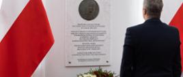 Wojewoda kujawsko-pomorski stoi pod tablicą upamiętniająca prezydenta Lecha Kaczyńskiego w otoczeniu dwóch biało-czerwonych flag