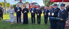W kształcie litery L stoją zaproszeni goście, wszyscy trzymają w rękach otrzymane pamiątkowe tabliczki, z tyłu widać samochód strażacki