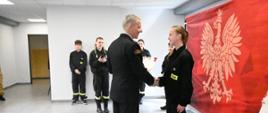 Strażak w mundurze wyjściowym gratuluje dziewczynie i wręcza dyplom. W prawej części zdjęcia czerwony baner PSP. W tle przed białą ścianą stoją inni uczestnicy turnieju. 