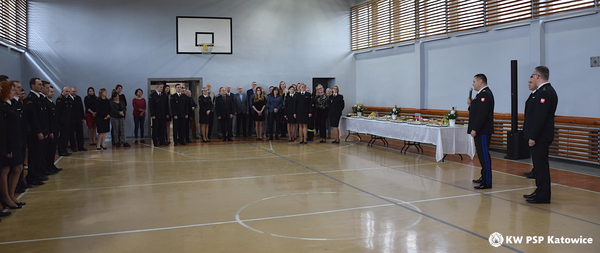 Na zdjęciu sala gimnastyczna w Komendzie Wojewódzkiej PSP w Katowicach. Po prawej stronie stoi w mundurze wyjściowym z mikrofonem śląski komendant wojewódzki nadbryg. Jacek Kleszczewski oraz zastępcy: st. bryg. Mirosław Synowiec i mł. bryg. Arkadiusz Krzemiński. Obok stół ze świątecznym nakryciem. Na wprost i po lewej stronie stoją funkcjonariusze i pracownicy cywilni komendy oraz zaproszeni goście.