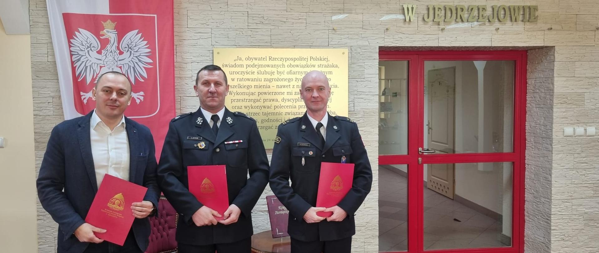 Na zdjęciu widnieje trzech mężczyzn znajdujących się na Sali Tradycji Komendy Powiatowej PSP w Jędrzejowie. Cała trójka trzyma w rękach podpisane porozumienie dotyczące włączenie jednostki Ochotniczej Straży Pożarnej w Sprowie w struktury krajowego systemu ratowniczo-gaśniczego.
