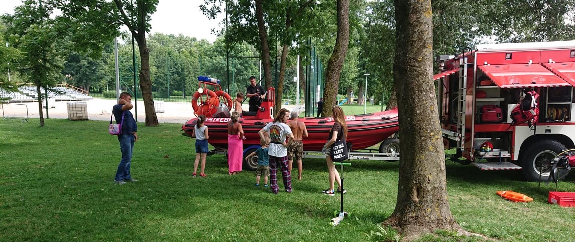 Zdjęcie przedstawia samochód strażacki, łódź, strażaków i ludzi oglądających łódź 