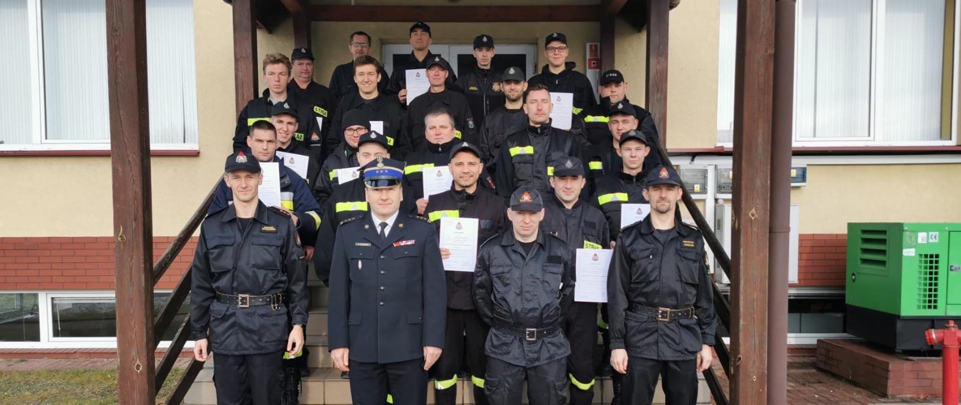 Na schodach zewnętrznych, zadaszonych, prowadzących do drzwi wejściowych do komendy, kolejno na stopniach ku górze stoją druhowie OSP, którzy ukończyli szkolenie podstawowe, ubrani w czarne umundurowanie koszarowe, trzymający w dłoniach zaświadczenia o ukończeniu szkolenia. Na samym dole, w pierwszym rzędzie stoi Komendant komendy oraz komisja egzaminacyjna, składająca się z oficerów Komendy Powiatowej Straży pożarnej w Rawie Mazowieckiej.