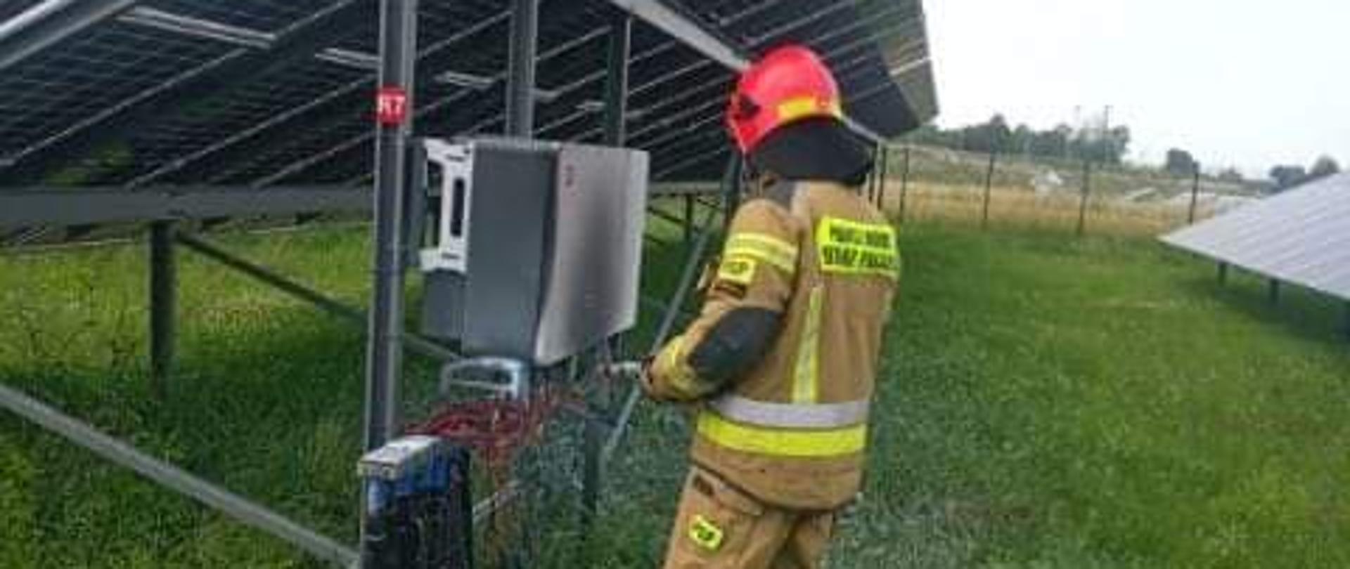 Strażak w ubraniu specjalnym stoi tyłem przed spalonym falownikiem na fermie fotowoltaicznej w miejscowości Skorków. Sprawdza kamerą termowizyjną ewentualne zarzewia ognia. 