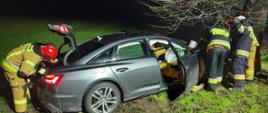 Widać uszkodzony samochód od strony pasażera z otwartymi drzwiami blisko drzewo, w pobliżu kilku strażaków
