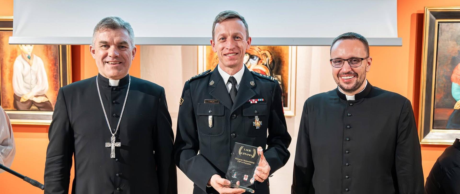 Komendant wraz z dyrektorem Caritas i Arcybiskupem stoją na zdjęciu, po wręczeniu statuetki - wyróżnienia.