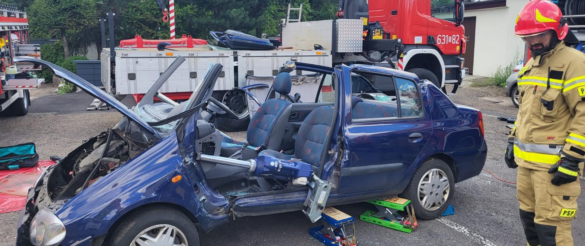 Granatowe auto bez drzwi i dachu, otwarta przednia maska. po prawej stronie stoi strażak 