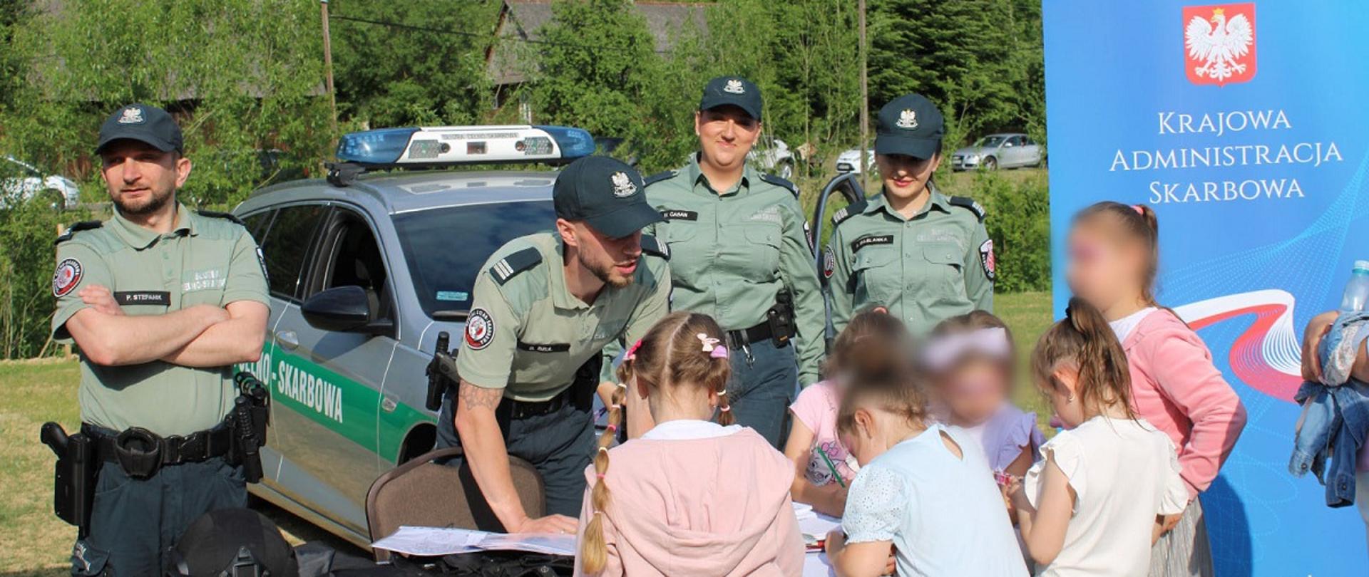 Piknik dla dzieci. Przy stanowisku podkarpackiej KAS dzieci oglądają sprzęt służbowy. Obok Stoją funkcjonariusze SCS . W tle radiowóz i baner KAS.