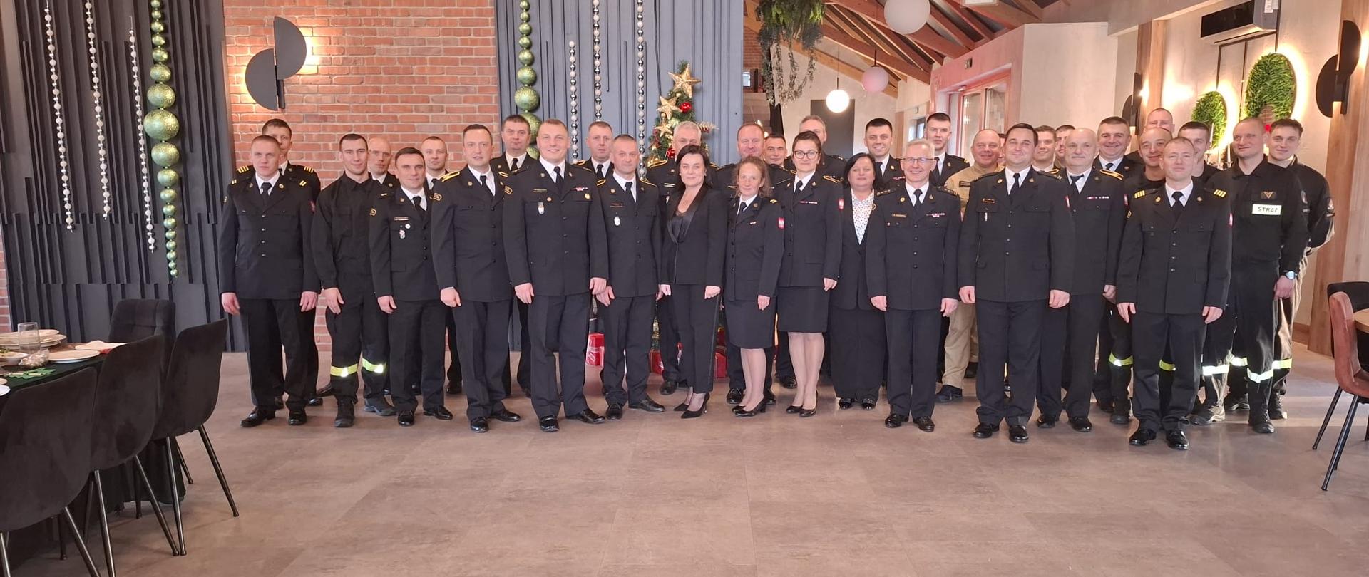 Grupowe zdjęcie wszystkich uczestników wigilii, którzy pozują w dwóch szeregach na tle ściany z cegieł, za nimi widać kontur choinki, po środku pozujących stoi Komendant Powiatowy PSP w Brzezinach starszy brygadier Mariusz Kaczmarski
