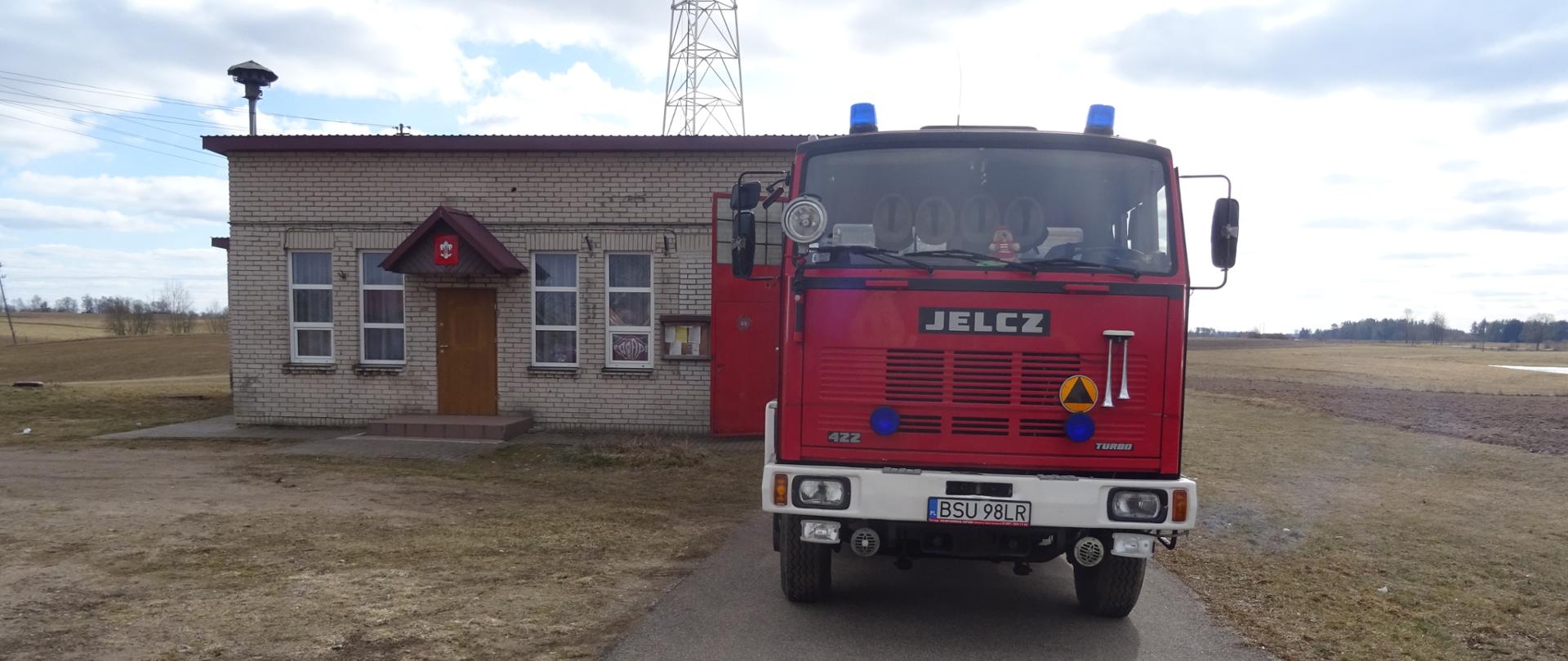 Na zdjęciu znajduje się ciężki samochód pożarniczy marki Jelcz w tle remiza OSP w Sidorach budynek parterowy o jasno szarych ścianach wokół pola