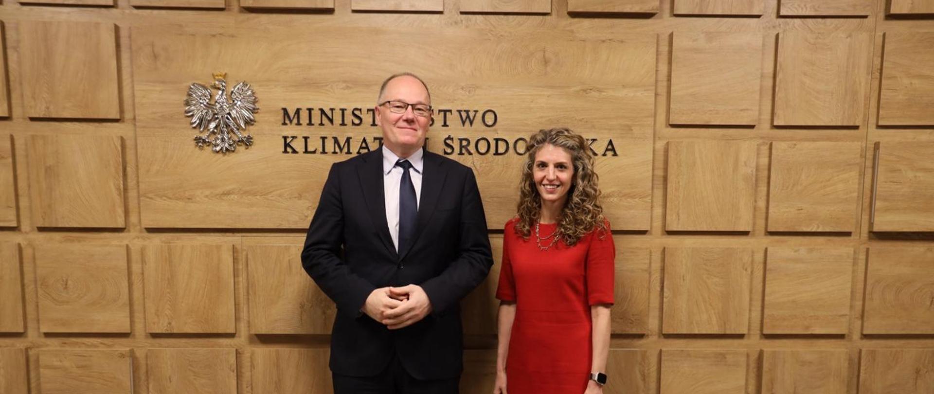 Deputy Minister Krzysztof Galos, Chief National Geologist, during a meeting with Stephanie Holmes, Deputy Chief of Mission at the U.S. Embassy in Poland. The photograph shows the two officials standing side by side and posing for a joint photo. The words “Ministry of Climate and Environment” can be seen in the background.