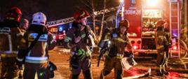 Funkcjonariusze w ubraniach specjalnych w trakcie prowadzenia działań gaśniczych, w tle czerwony samochód gaśniczy